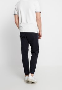 Mies, joka käyttää valkoista lyhythihaista pooloa, mustia jogger-housuja ja valkoisia Veja-tenniskenkiä, seisoo selkä kameraan päin yksinkertaista valkoista taustaa vasten.