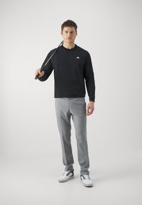 Svart långärmad skjorta, grå byxor och vita sneakers. Modellen håller en golfklubba, med en enkel och stilren design.