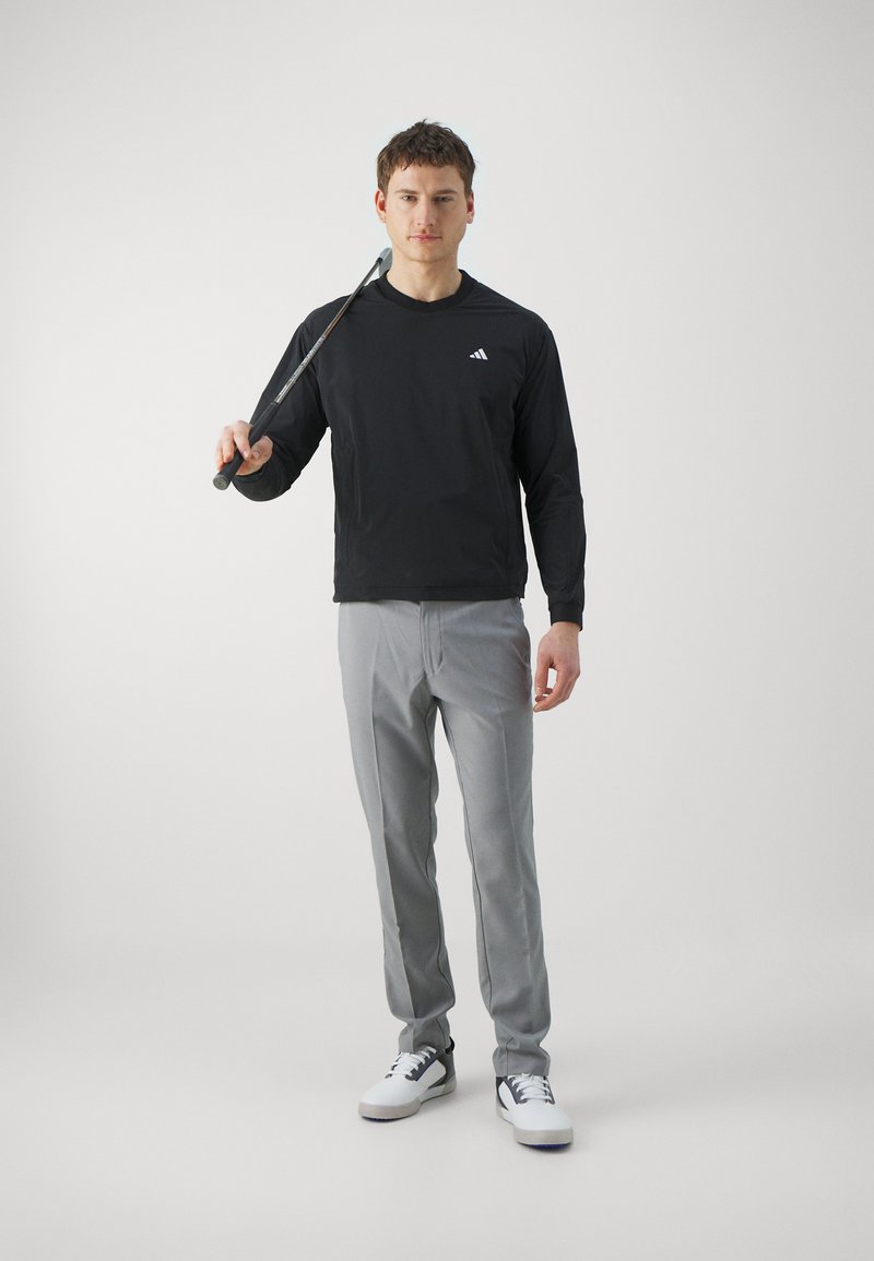 Schwarzes Langarmshirt, graue Hose und weiße Sneakers. Das Model hält einen Golfschläger, mit einem schlichten und eleganten Design.