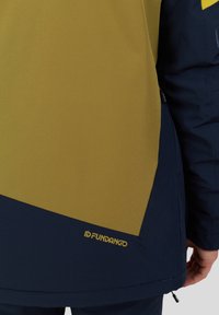 Giacca gialla e blu con un design geometrico, texture liscia e logo visibile sul bordo inferiore.