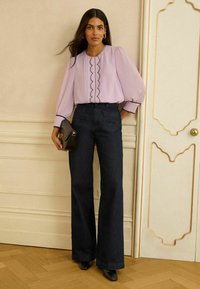 Laventelinvärinen paita pussihihoilla ja mustilla siksak-reunakuvioilla, yhdistetty korkeavyötäröisiin tummansinisiin leveälahkeisiin farkkuihin ja mustiin teräväkärkisiin kenkiin.