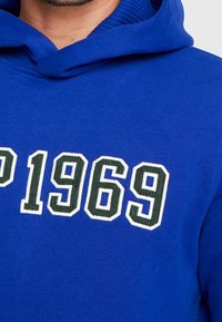 Sudadera con capucha azul que presenta letras en blanco y verde oscuro "AP 1969"; textura suave y interior acanalado debajo de la capucha.