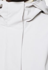 Světlounce šedá nepromokavá bunda s vysokým límcem, jemnými švy a hladkým matným povrchem. Má nastavitelnou kapuci se stahovací šňůrkou.