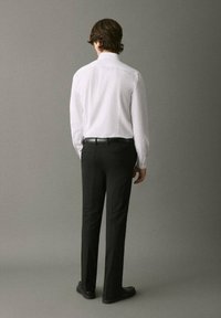 Camisa blanca de manga larga con cuello puntiagudo, combinada con pantalones negros a medida y zapatos negros. Tomada desde la parte trasera contra un fondo gris.