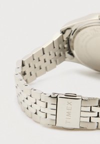 Sudraba metāla pulksteņa siksna ar sprādzi, uz kuras ir iegravēts "TIMEX", uz baltas fona, parādot daļu pulksteņa aizmugures.