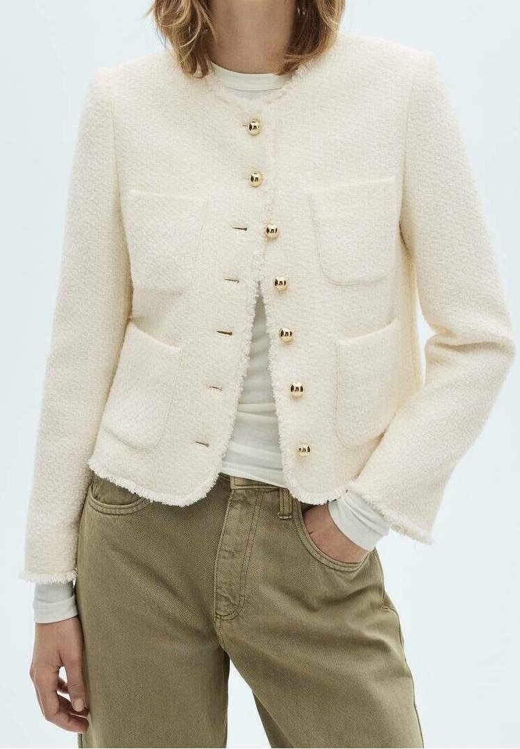 Femme portant une veste crème texturée avec des boutons dorés, un haut à manches longues blanc et un pantalon vert olive, la main dans la poche.