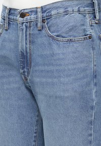 Ανοιχτό μπλε τζιν παντελόνι φτιαγμένο από denim, φορεμένο στη μέση με ορατό μπροστινό κουμπί, θηλιές για ζώνη, τσέπες και πορτοκαλί ραφές.