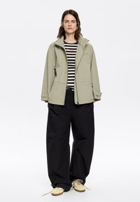Giacca a vento verde chiaro con cappuccio e due tasche frontali, indossata sopra una maglietta a righe bianche e nere. Abbinata a pantaloni neri larghi e scarpe chiare.