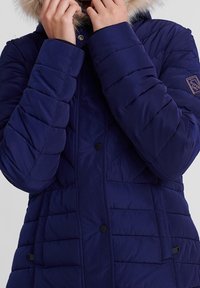 Marineblå polstret jakke med dyretrimsbeklædning på hætten, med skrå quiltet design, trykknapper og sidelommer. Holdbart stof.