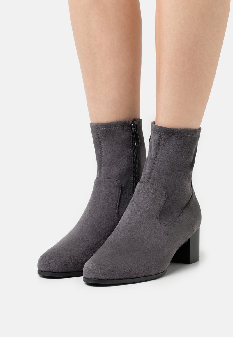 Bottes cheville en daim gris avec talons carrés et fermetures éclair latérales portées sur des jambes nues contre un fond blanc uni.