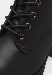 Gros plan d'une botte en cuir noir avec des lacets noirs glissés à travers des œillets métalliques argentés et une couture visible sur le côté.