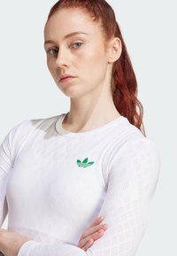 White long-sleeve athletic shirt with a textured pattern, featuring a small green Adidas logo on the chest. Smooth, fitted design.