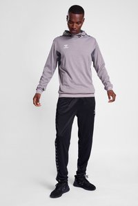Felpa grigia con cappuccio e logo, abbinata a pantaloni da tuta neri con motivo a chevron laterale. Realizzati in tessuto leggero.
