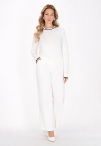 Top alb cu maneci lungi si decolteu negru, asortat cu pantaloni albi cu croiala larga. Modelul poarta de asemenea pantofi albi si cercei mari, stand in fata unui fundal simplu.