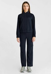 Marineblauw trainingspak met een jas met hoge kraag en cargobroek. De jas heeft een subtiel logo; de broek heeft zijzakken. Glad stof, comfortabele pasvorm.