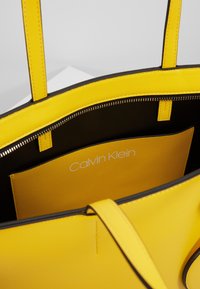 Żółta skórzana torba tote z czarnym wnętrzem i zamkiem błyskawicznym, pokazująca wewnętrzną żółtą kieszeń z widocznym białym logo Calvin Klein.