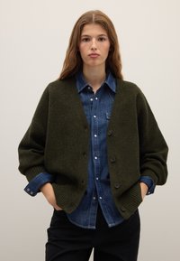 Olivgrüne Wollstrickjacke mit lockerem Schnitt, gerippten Bündchen und fünf Knöpfen, getragen über einem Jeanshemd mit Knopfleiste.