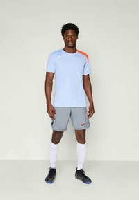 T-shirt de sport bleu clair avec des accents orange, shorts gris et chaussures de football noires, complété par des chaussettes blanches jusqu'aux genoux. Marque Nike visible.