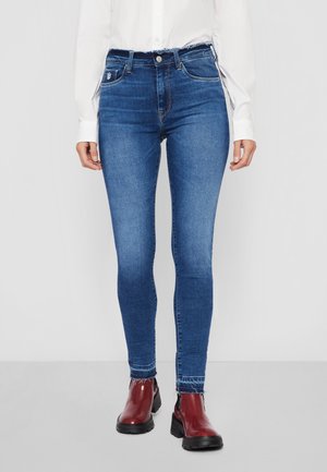 Nainen, jolla on siniset kapeat farkut, joissa on rispaantuneet reunat, yhdistettynä tummanpunaisiin nilkkureihin ja valkoiseen pitkähihainen paitaan, seisomassa yksivärisen taustan edessä.