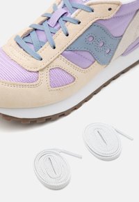 Sneakers med en beige mockaovandel, lavendelfärgade nätinslag och logotyp i grått läder. Inkluderar två vita skosnören mot en neutral bakgrund.