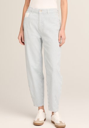 Person trägt hellblau-weiße vertikal gestreifte Hosen mit Taschen und weiße Turnschuhe vor neutralem Hintergrund.