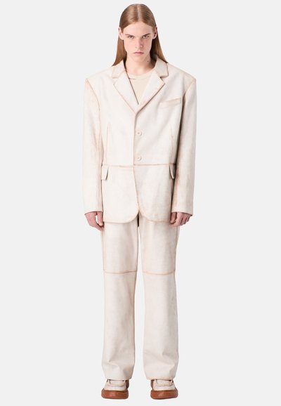 Beige maßgeschneiderter Anzug mit einem einreihigen Sakko mit Reverskragen und zwei Knöpfen, kombiniert mit weit geschnittenen Hosen. Strukturierter Stoff.