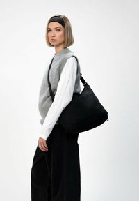 Schwarze Umhängetasche mit glatter Oberfläche, verstellbarem Riemen und dezentem Branding, getragen über einem grauen Pullover und einem weißen Hemd, kombiniert mit schwarzen Hosen.