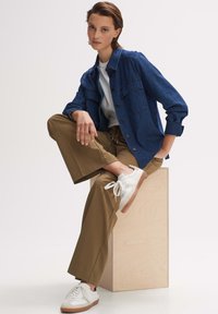 Denim jacket in dark blue with button front and two chest pockets, paired with brown wide-leg trousers and white sneakers with beige accents.