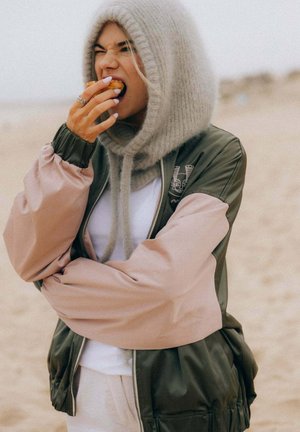Young woman wearing hooded knit hat and two-tone jacket biting into a pastry while standing on a sandy beach.