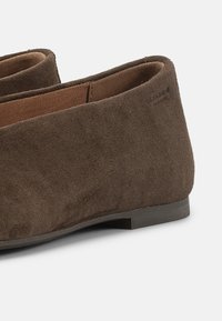 Ζευγάρι καφέ μοκασίνια από σουέτ με χαμηλά τακούνια και ελαστικές πλαϊνές επιφάνειες σε λευκό φόντο.