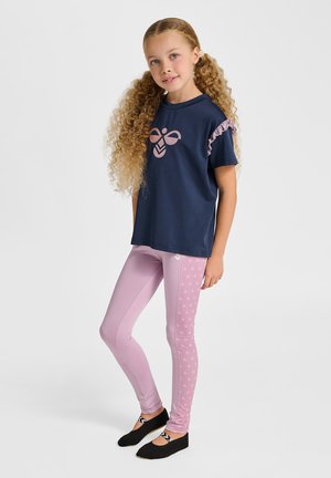 Ung pige med krøllet hår iført en mørkeblå skjorte med pink logo, lavendel leggings med mønstre på siderne og sorte slip-on sko.