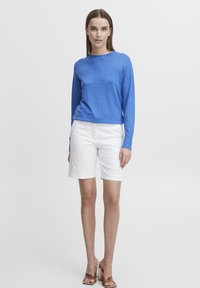 Haut à manches longues bleu en matière tricotée douce, associé à un short blanc arrivant aux genoux et des sandales à talons bruns. Design simple et décontracté.