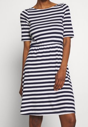 Femme portant une robe blanche mi-longue avec des rayures horizontales bleu marine, manches courtes et taille cintrée, debout devant un fond uni.