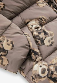 Tessuto marrone trapuntato con motivi di orsetti di peluche ricamati in diverse tonalità di beige e marrone scuro.