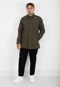 Giacca impermeabile marrone con colletto, tasche frontali e logo sul petto, abbinata a pantaloni neri e sneakers bianche.