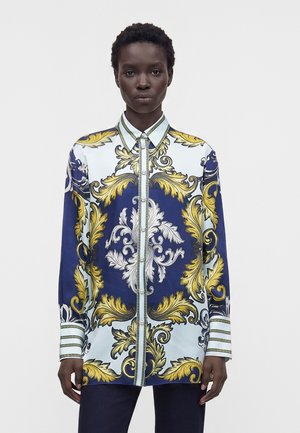Personne portant une chemise à manches longues boutonnée avec un motif floral orné jaune et blanc sur un fond bleu et bleu clair.