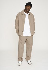 Beige Baumwolljacke mit Knopfleiste, weißem T-Shirt, passenden beigen Hosen und schwarzer Mütze. Die Sneakers haben schwarze Streifen auf weißem Grund.