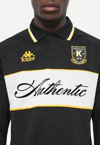 Polo a maniche lunghe nera con una striscia gialla e bianca. Presenta il logo Kappa e la scritta "Authentic" in corsivo, con colletto polo.