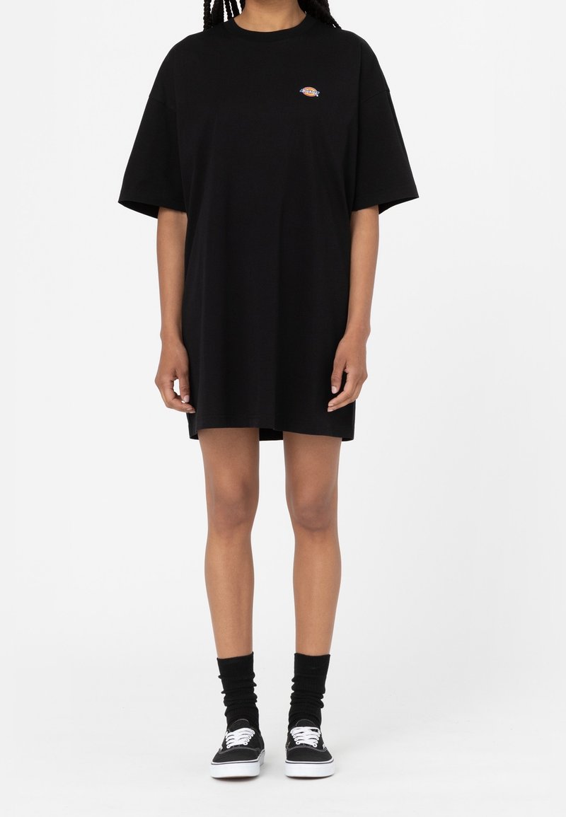 Robe t-shirt oversize noire à manches courtes, avec un petit logo coloré sur la poitrine. Portée avec des chaussettes noires et des chaussures en toile.