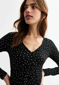 Top negro con mangas largas, escote en V, con una tela texturizada y un patrón de lunares blancos. Los detalles incluyen acentos de costura en la parte delantera.