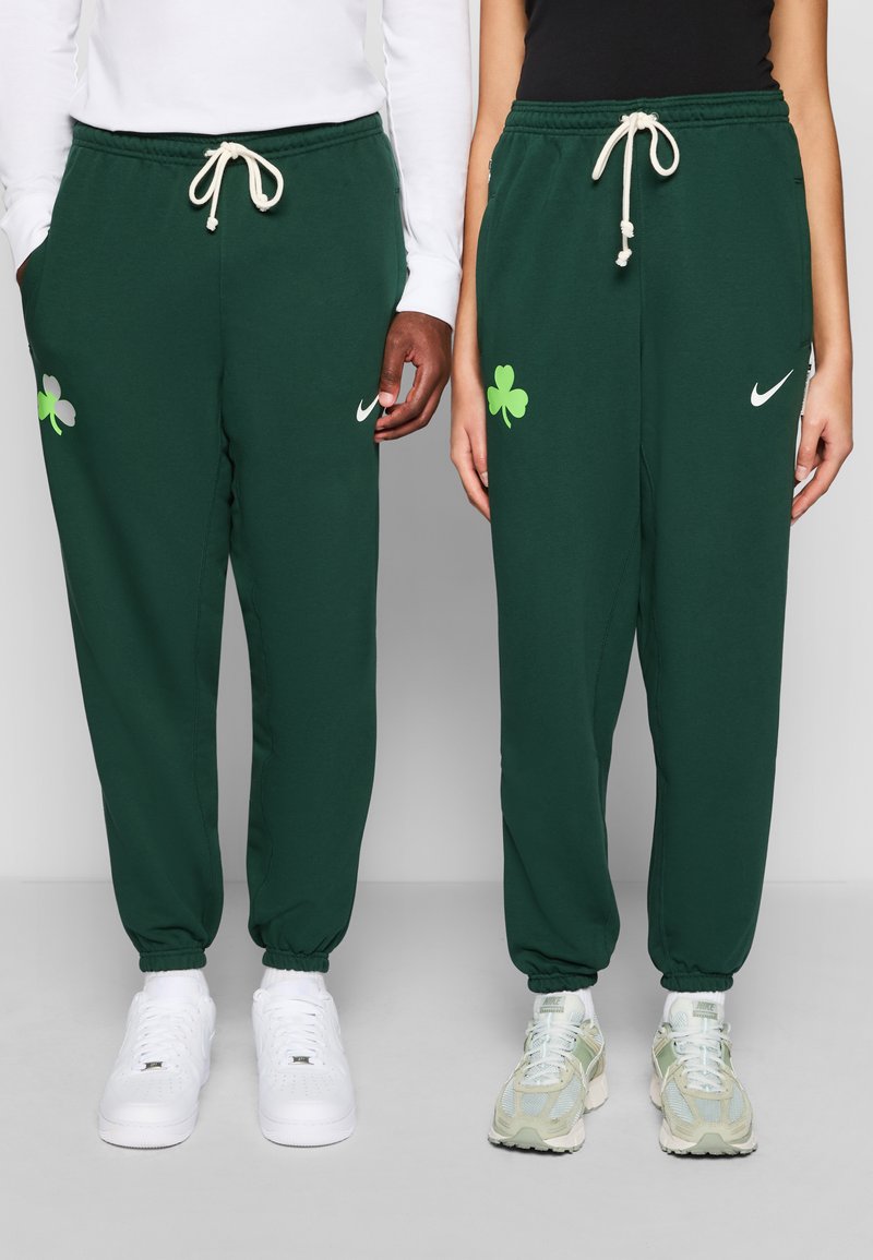 Calças de treino verdes feitas de uma mistura de algodão, com um cordão branco e bolsos laterais. À esquerda, possui um logo branco da Nike, enquanto à direita apresenta um detalhe verde de trevo.