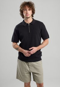 Black ribbed polo shirt with a zippered collar and short sleeves, paired with light beige shorts. Simple design with subtle texture.