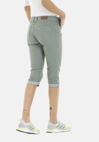 Pantalons cropped verts clair avec des poignets roulés, dotés de poches arrière et d'une finition texturée, associés à des baskets grises rehaussées d'accents jaunes.