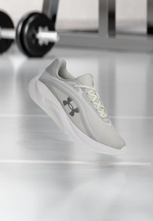 Lysegrå Under Armour løbesko med hvid sål svæver over et reflekterende fitnessgulv, håndvægtssæt sløret i baggrunden.