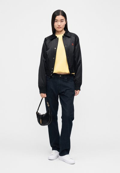 Čierna saténová bomber bunda s golierom, žltý bavlnený sveter, tmavé denimové džínsy a malá čierna kabelka. Oblečenie dopĺňajú biele tenisky.