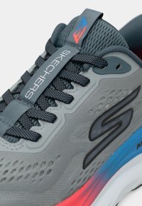 Gros plan d'une chaussure de course Skechers grise avec des lacets noirs, une semelle bleue et rouge, et des trous de ventilation sur le tissu supérieur.