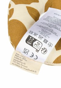 Medvilninė pagalvėlė su abstrakčiu garstyčių geltonos ir kreminės spalvų lapų raštu. Prie jos prikabinta priežiūros etiketė su skalbimo simboliais ir saugumo sertifikatais.