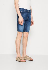 Shorts in denim blu medium con taglio dritto, orli risvoltati e leggera scoloritura. Indossati con sandali beige senza cinturino.