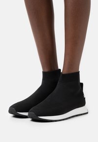 Svarta stickade fotledsstövlar med ribbad krage och vit sula. Har en strukturerad yta och en liten logotyp på sidan.