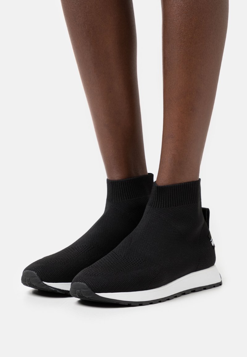 Svarta stickade fotledsstövlar med ribbad krage och vit sula. Har en strukturerad yta och en liten logotyp på sidan.